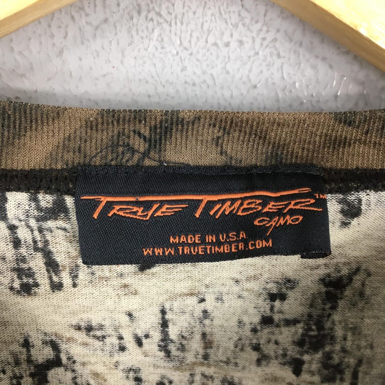 May include: Close-up of a camouflage pattern fabric with a black label. The label reads "True Timber Camo" in orange lettering, with "Made in U.S.A." and a website address below. The fabric has a brown and beige color scheme.