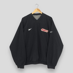 Könnte beinhalten: Eine schwarze langärmelige Windjacke zum Überziehen mit V-Ausschnitt. Auf der Vorderseite befindet sich ein weißes Reebok-Logo auf der rechten Brust und ein rot-weißes 49ers-Teamlogo auf der linken Brust. Die Jacke hat lange Ärmel mit elastischen Bündchen.