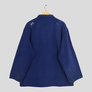 Peut inclure: Veste de travail bleu marine vintage &agrave; manches longues, suspendue &agrave; un cintre en bois. La veste pr&eacute;sente des signes d'usure, avec des &eacute;claboussures de peinture et des imperfections mineures. Le col est de style classique.