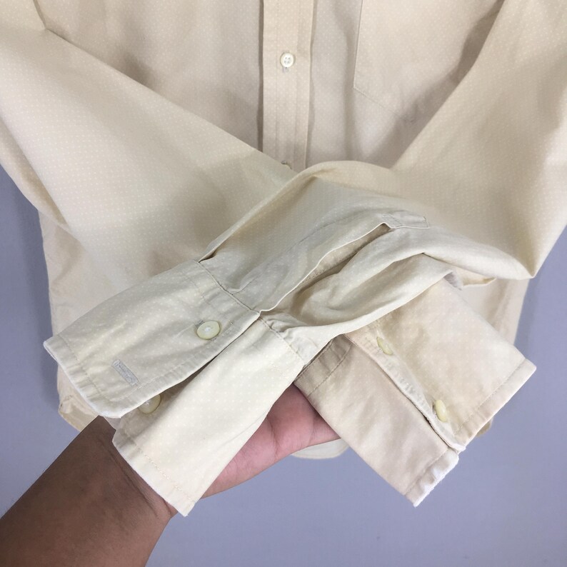 Pode incluir: Uma camisa de manga comprida bege claro com um padr&atilde;o sutil. A camisa tem um punho de bot&atilde;o &uacute;nico e um bolso no peito esquerdo.