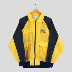 Puede incluir: Chaqueta amarilla y azul marino con cuello y cremallera completa. La chaqueta tiene dos bolsillos delanteros y un pequeño parche que dice "Benetton Formula". Las mangas y el dobladillo son de color azul marino.