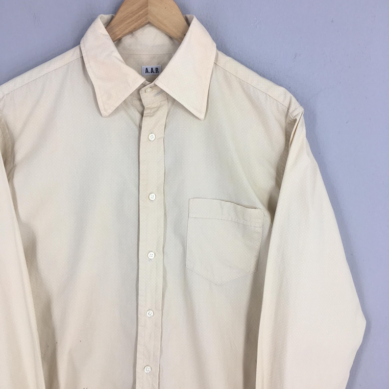 Pode incluir: Uma camisa de bot&otilde;es de manga comprida, de cor creme, com um bolso no peito e uma pequena etiqueta "A.A.R" na frente.