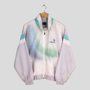 May include: A vintage, full-zip track jacket in a light purple and white color scheme. The jacket features a spray-paint design in shades of blue and green. Accents include teal stripes on the sleeves and collar. The Sergio Tacchini logo is on the chest.