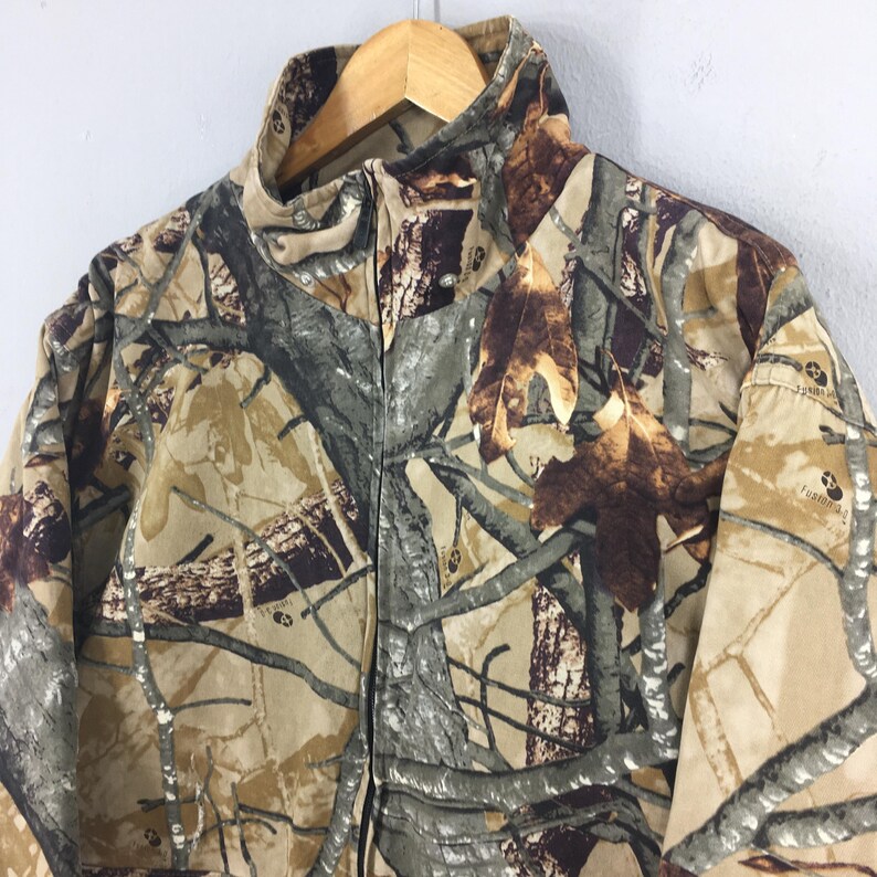 May include: A camouflage jacket with a full zipper closure. The jacket features a pattern of brown, tan, and green, resembling a forest environment. The jacket is hanging on a wooden hanger.