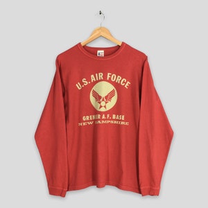 May include: Red long-sleeve shirt with "U.S. AIR FORCE" printed in cream-colored letters and a circular emblem. The emblem features a pair of wings and a star. Below the emblem, the text reads "GRENIER A.F. BASE NEW HAMPSHIRE".