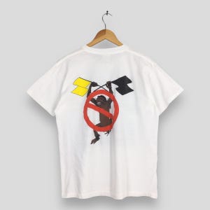 May include: White t-shirt featuring a graphic of a brown monkey inside a red circle with a line through it. The monkey is holding yellow and black flags. The shirt is hanging on a wooden hanger.