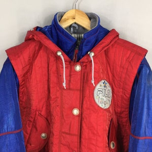 May include: A vintage red and blue Fila jacket with a hood. The jacket features a button-up front, two pockets, and the Fila Italy logo on the chest. The jacket is hanging on a wooden hanger.
