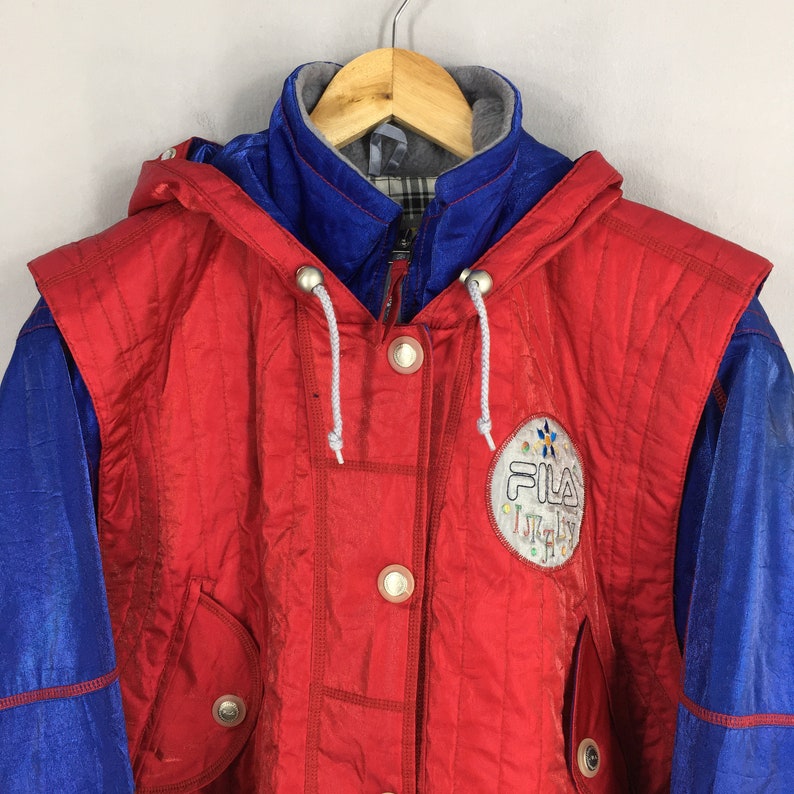 May include: A vintage red and blue Fila jacket with a hood. The jacket features a button-up front, two pockets, and the Fila Italy logo on the chest. The jacket is hanging on a wooden hanger.