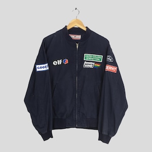 Puede incluir: Chaqueta bomber azul marino con varios parches, incluyendo "GOOD YEAR", "elf" y "Benetton Formula Racing Team". La chaqueta tiene cierre de cremallera y puños y dobladillo acanalados. Una chaqueta de carreras vintage.