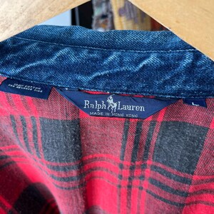 May include: A Ralph Lauren shirt featuring a dark blue denim collar and a red and black plaid flannel body. The interior neck label displays the brand name, a polo player logo, and the text Made in Hong Kong. A secondary tag indicates 100% cotton.