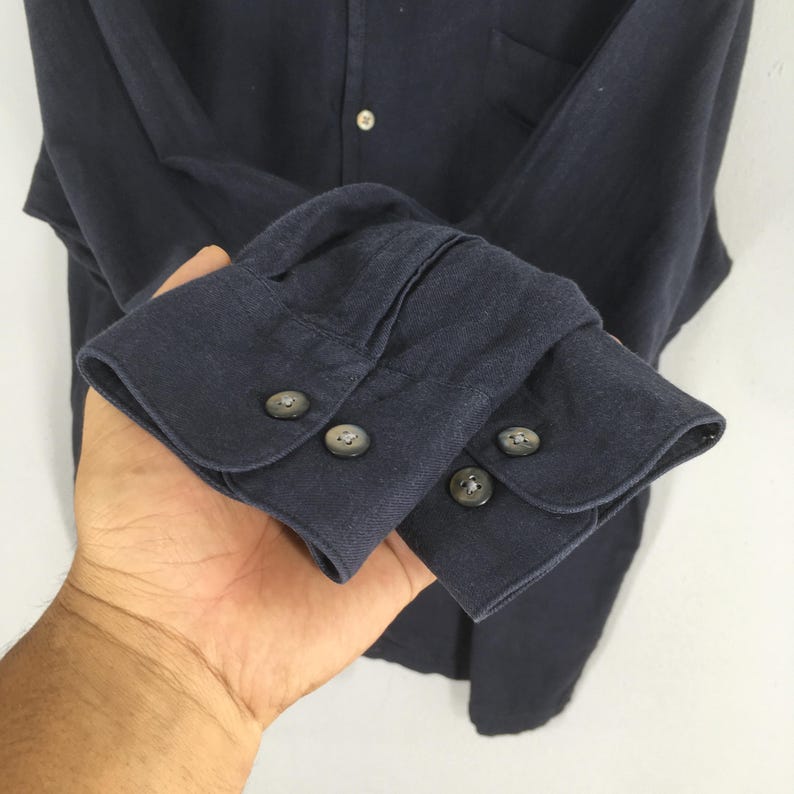 Puede incluir: Primer plano de una camisa azul marino de manga larga. La camisa presenta un dise&ntilde;o abotonado con cuello cl&aacute;sico y pu&ntilde;os abotonados. Los pu&ntilde;os tienen tres botones grises cada uno. La tela parece ser un material suave y tejido.