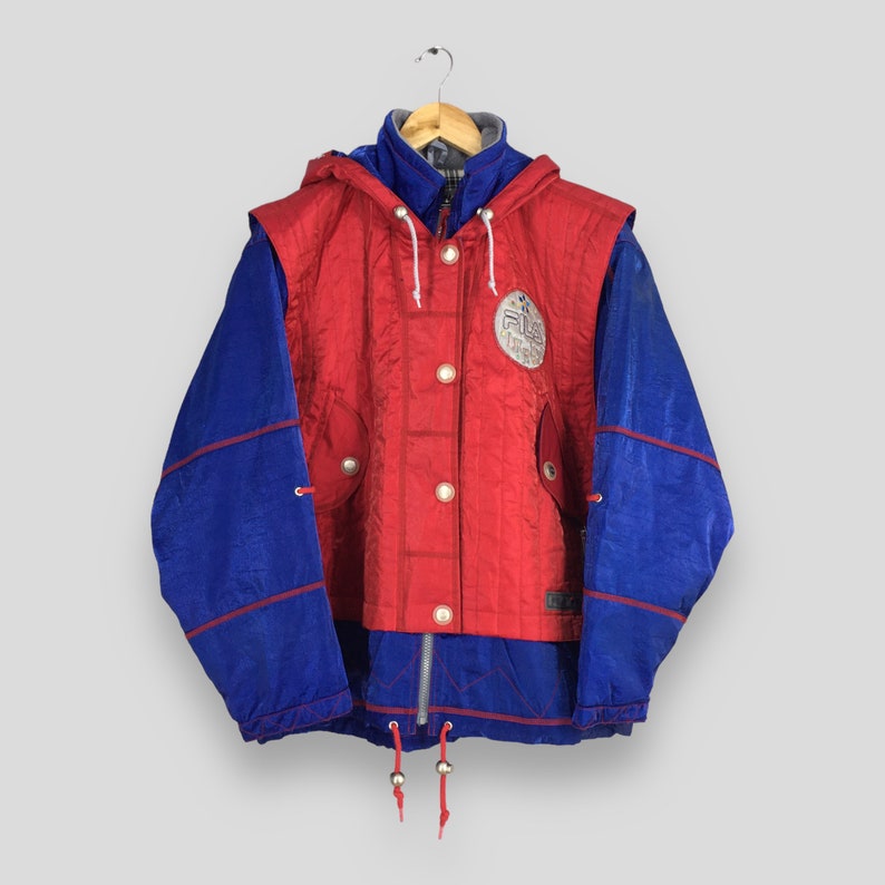 May include: A vintage red and blue Fila jacket with a hood, button-up front, and a logo patch. The jacket features a color-block design with blue sleeves and red panels. It is hanging on a wooden hanger.