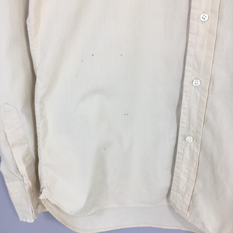 Pode incluir: Uma camisa de bot&otilde;es de manga comprida de cor creme com um padr&atilde;o sutil de bolinhas brancas. A camisa tem alguns pequenos furos.