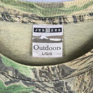 May include: Close-up of a camouflage-patterned t-shirt. The shirt features a tag with the brand name "JERZEES" and the word "Outdoors" printed in black. The shirt is a blend of green, brown, and tan colors.
