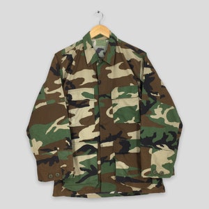 Puede incluir: Chaqueta con estampado de camuflaje con mangas largas, cuello y cuatro bolsillos delanteros. La chaqueta es en tonos verdes, marrones y beige. Cuelga de una percha de madera.
