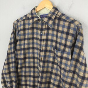 K&ouml;nnte beinhalten: Ein lang&auml;rmeliges Flanellhemd mit Kn&ouml;pfen in einem Karomuster aus Blau, Beige und Marine. Das Hemd hat eine einzelne Brusttasche und einen Kragen. Das Hemd h&auml;ngt an einem Holzb&uuml;gel.