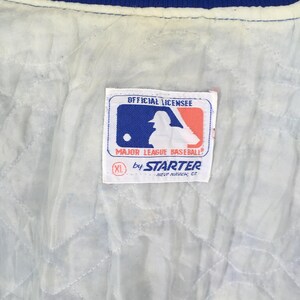 May include: A white quilted jacket lining featuring a rectangular tag with the Major League Baseball logo, the text OFFICIAL LICENSEE, MAJOR LEAGUE BASEBALL, by STARTER, NEW HAVEN, CT, and a red circle containing the size XL.