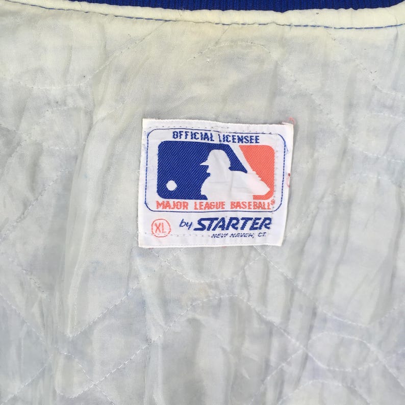 May include: A white quilted jacket lining featuring a rectangular tag with the Major League Baseball logo, the text OFFICIAL LICENSEE, MAJOR LEAGUE BASEBALL, by STARTER, NEW HAVEN, CT, and a red circle containing the size XL.