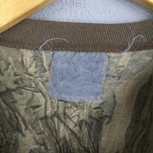 May include: Close-up of a camouflage pattern t-shirt. The shirt features a brown ribbed neckline and a light blue tag. The camouflage design incorporates shades of green, brown, and tan, creating a textured appearance. The shirt is hanging on a wooden hanger.