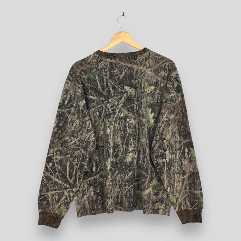 May include: Long-sleeved camouflage shirt with a brown collar and cuffs. The shirt features a detailed pattern of branches, leaves, and bark in shades of brown, green, and tan. The shirt is hanging on a wooden hanger.