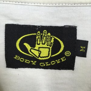 Peut inclure: &Eacute;tiquette noire avec les mots "BODY GLOVE" en jaune et un logo de main jaune &agrave; l'int&eacute;rieur d'un ovale. Une petite &eacute;tiquette noire avec la lettre "M" est attach&eacute;e. L'&eacute;tiquette est sur un tissu blanc.