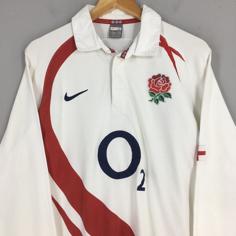 Peut inclure: Maillot de rugby blanc &agrave; manches longues avec des d&eacute;tails rouges et un &eacute;cusson de rose. Comprend un logo "O2" bleu marine et le logo Nike. Le maillot a un col et est suspendu &agrave; un cintre en bois.