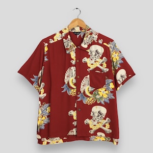 Puede incluir: Camisa de manga corta con botones en un tono rojo oscuro, adornada con un patrón repetido de calaveras, huesos cruzados y elementos florales. La camisa presenta un cuello clásico y un bolsillo en el pecho, creando un diseño audaz y llamativo.