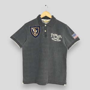 Puede incluir: Una camiseta polo gris con un parche de un tigre volador en el pecho izquierdo y el texto "Flying Tiger's American Volunteer Group" en el pecho derecho. La camiseta también tiene un pequeño parche de la bandera estadounidense en la manga derecha.