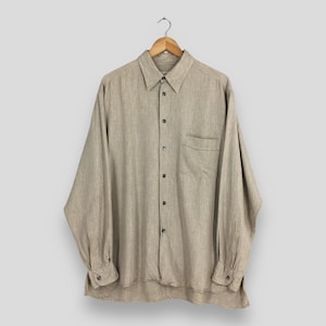 May include: A long-sleeved, button-down shirt in a neutral beige color, hanging on a wooden hanger. The shirt features a chest pocket and buttoned cuffs. The fabric appears to be a linen or linen blend.