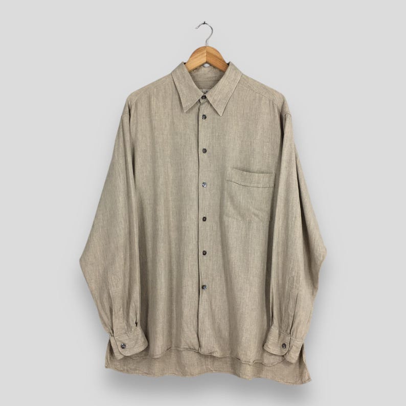 May include: A long-sleeved, button-down shirt in a neutral beige color, hanging on a wooden hanger. The shirt features a chest pocket and buttoned cuffs. The fabric appears to be a linen or linen blend.
