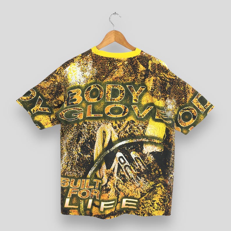Peut inclure: T-shirt vintage Body Glove avec col jaune et imprim&eacute; int&eacute;gral. Le design pr&eacute;sente les mots "BODY GLOVE" et "BUILT FOR LIFE" dans une police audacieuse et textur&eacute;e, avec un fond de motifs et d'images abstraits.