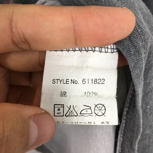 May include: A gray fabric tag with the text "STYLE No. 611822" and "綿 100%" printed in black. The tag also features laundry symbols. The tag is attached to a gray garment.