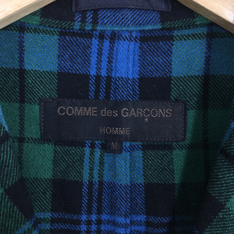K&ouml;nnte beinhalten: Ein schwarzes und gr&uuml;nes kariertes Hemd mit einem schwarzen Etikett, auf dem "COMME des GARCONS HOMME" und "M" in wei&szlig; steht.