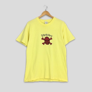 以下が含まれることがあります： 黄色い半袖Tシャツに、赤いドクロとクロスボーンのグラフィックと、黒い「Thrasher」の文字がプリントされています。