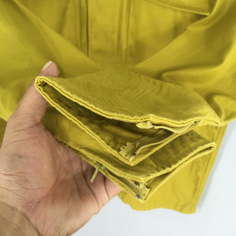Pode incluir: Close-up de uma pe&ccedil;a de roupa amarelo mostarda, possivelmente um casaco ou cal&ccedil;as, com costuras e z&iacute;peres vis&iacute;veis. O tecido parece ser uma mistura resistente de algod&atilde;o ou linho. A pe&ccedil;a de roupa &eacute; segurada por uma m&atilde;o.