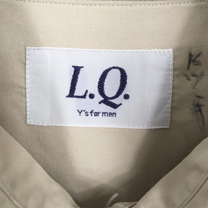 Peut inclure: Gros plan sur un col de chemise beige avec une &eacute;tiquette blanche. L'&eacute;tiquette affiche le texte "L.Q." en bleu fonc&eacute;, avec "Y's for men" imprim&eacute; en dessous. Le tissu de la chemise a une texture subtile.