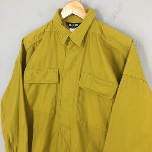 Pode incluir: Camisa mostarda de manga comprida com dois bolsos no peito e gola. A camisa est&aacute; pendurada em um cabide de madeira. A camisa tem uma frente abotoada e um pequeno logotipo na gola.