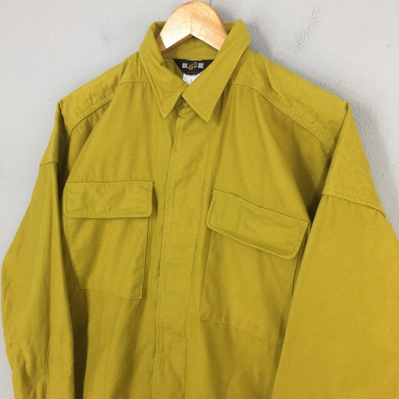 Pode incluir: Camisa mostarda de manga comprida com dois bolsos no peito e gola. A camisa est&aacute; pendurada em um cabide de madeira. A camisa tem uma frente abotoada e um pequeno logotipo na gola.