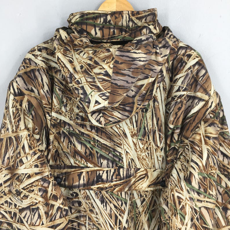 Puede incluir: Una chaqueta con capucha de camuflaje en tonos marrones, beige y verdes, dise&ntilde;ada para integrarse en entornos naturales. La chaqueta presenta una capucha completa, cierres de bot&oacute;n y un patr&oacute;n que se asemeja a juncos y follaje. La chaqueta est&aacute; colgada en una percha de madera.