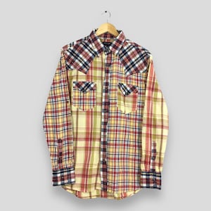 Puede incluir: Una camisa de manga larga con botones de cuadros amarillos, rojos y azules con dos bolsillos en el pecho. La camisa tiene un cuello de estilo western y está colgada de una percha de madera.