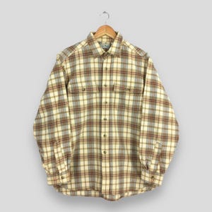 Puede incluir: Una camisa de franela a cuadros con botones y mangas largas en tonos beige, crema, marrón y gris. La camisa tiene dos bolsillos en el pecho y está colgada de una percha de madera. La etiqueta L.L. Bean es visible.