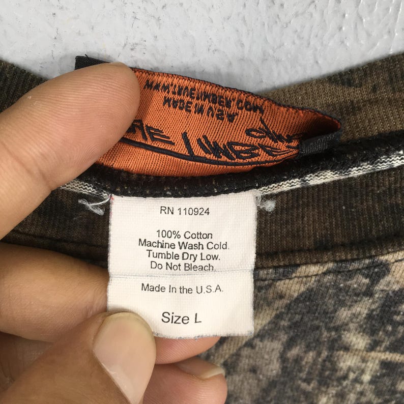 May include: Close-up of a clothing tag with the text "RN 110924", "100% Cotton", and "Made In the U.S.A.". The tag is white with black text and is attached to a camouflage-patterned fabric. The size is "L".