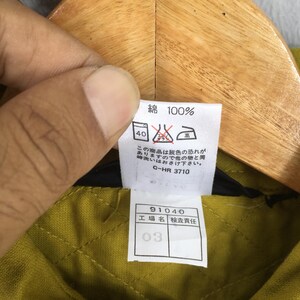 Pode incluir: Close-up de uma etiqueta de roupa com texto japon&ecirc;s e s&iacute;mbolos de cuidado. A etiqueta diz "綿 100%" (algod&atilde;o 100%) e inclui instru&ccedil;&otilde;es de lavagem. Abaixo, uma etiqueta menor exibe n&uacute;meros e caracteres japoneses.