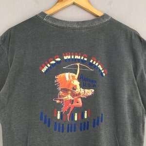 May include: Dark gray t-shirt with a graphic featuring the text "MISS WING DING" in red, white, and blue. The graphic includes a Native American woman with a bow and arrow, a vintage airplane, and bombs. The brand name "Avirex U.S.A." is also visible.