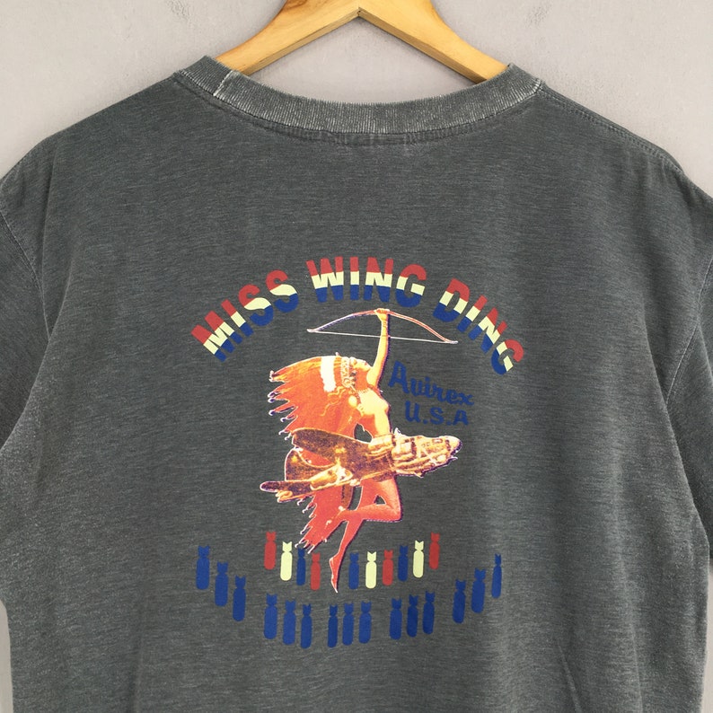 May include: Dark gray t-shirt with a graphic featuring the text "MISS WING DING" in red, white, and blue. The graphic includes a Native American woman with a bow and arrow, a vintage airplane, and bombs. The brand name "Avirex U.S.A." is also visible.
