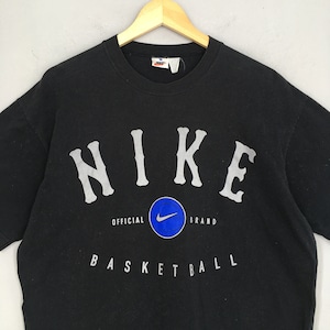 Może przedstawiać: Czarny T-shirt z białym logo "NIKE" i grafiką koszyk&oacute;wki. Na koszulce nadrukowany jest r&oacute;wnież tekst "OFFICIAL BRAND" i "BASKETBALL".