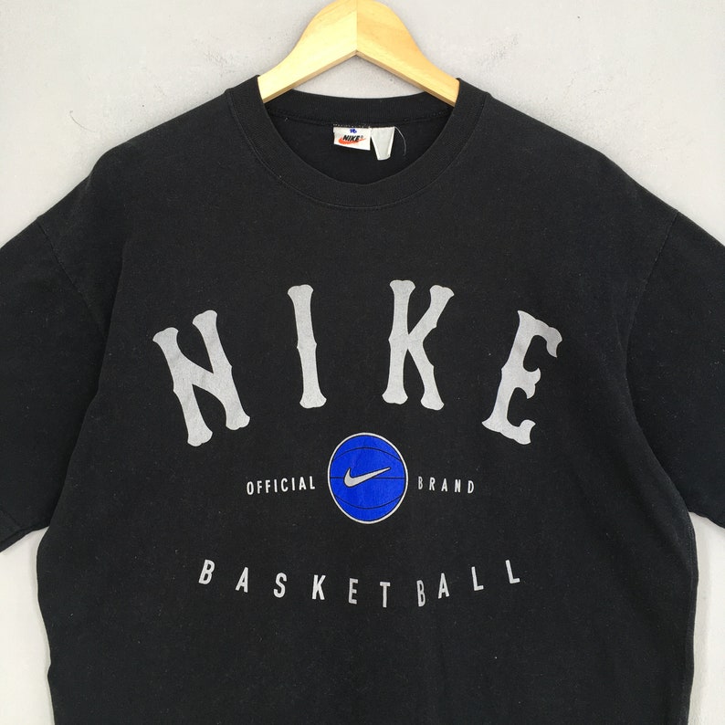 Może przedstawiać: Czarny T-shirt z białym logo "NIKE" i grafiką koszyk&oacute;wki. Na koszulce nadrukowany jest r&oacute;wnież tekst "OFFICIAL BRAND" i "BASKETBALL".