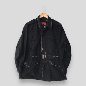 Könnte beinhalten: Eine schwarze Feldjacke mit Kragen, langen Ärmeln und vier Vordertaschen mit geknöpften Klappen. Die Jacke verfügt über Schulterklappen, einen Frontreißverschluss und sichtbare ausgefranste Kanten entlang der Nähte und Taschen.