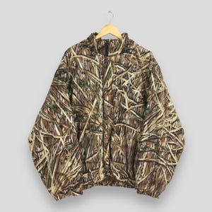Puede incluir: Chaqueta de camuflaje con cremallera completa, con un patr&oacute;n marr&oacute;n, beige y verde, colgada de una percha de madera. La chaqueta presenta un cuello alto, un peque&ntilde;o logotipo en el pecho y un dobladillo con cord&oacute;n. El dise&ntilde;o de camuflaje recuerda a juncos y hierbas.