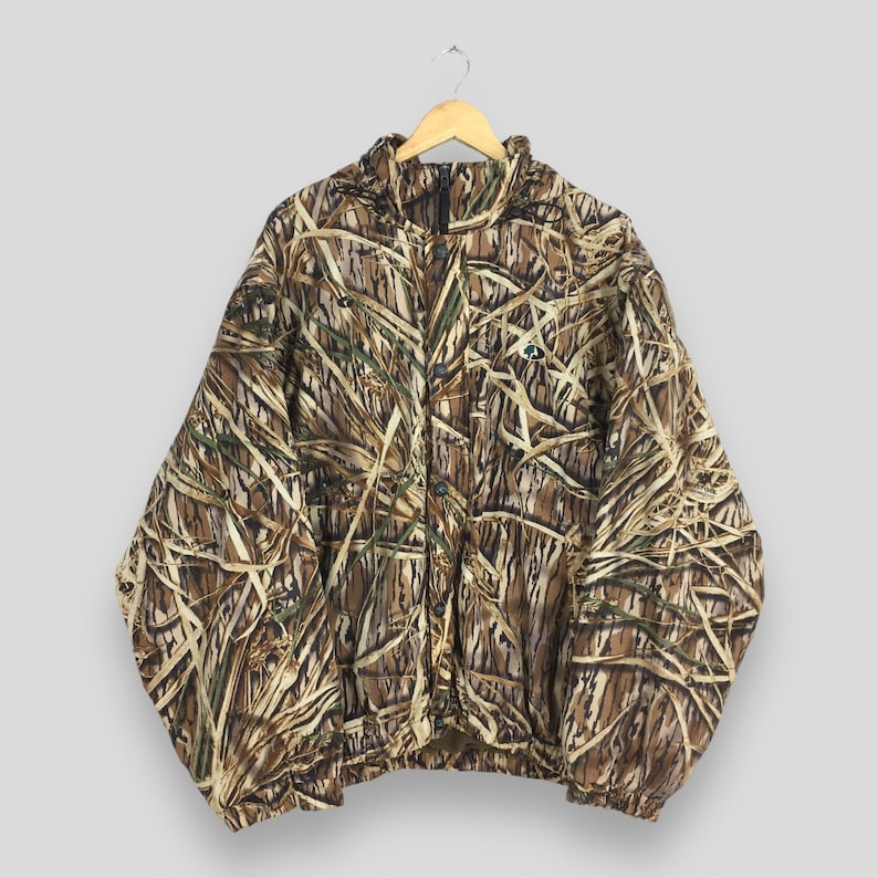 Puede incluir: Chaqueta de camuflaje con cremallera completa, con un patr&oacute;n marr&oacute;n, beige y verde, colgada de una percha de madera. La chaqueta presenta un cuello alto, un peque&ntilde;o logotipo en el pecho y un dobladillo con cord&oacute;n. El dise&ntilde;o de camuflaje recuerda a juncos y hierbas.